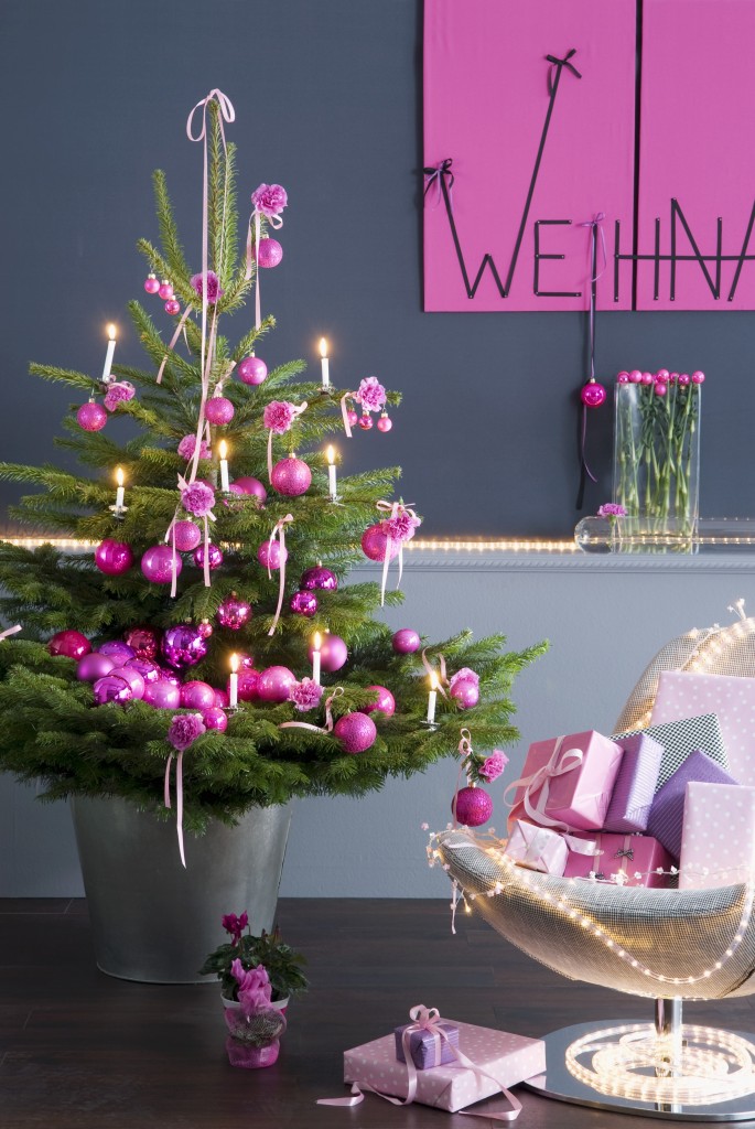 Geschmückter Weihnachtsbaum mit pinkfarbenen Kugeln neben Sessel mit Geschenken vor grauer Wand und Weihnachtsgruss an pinkfaben